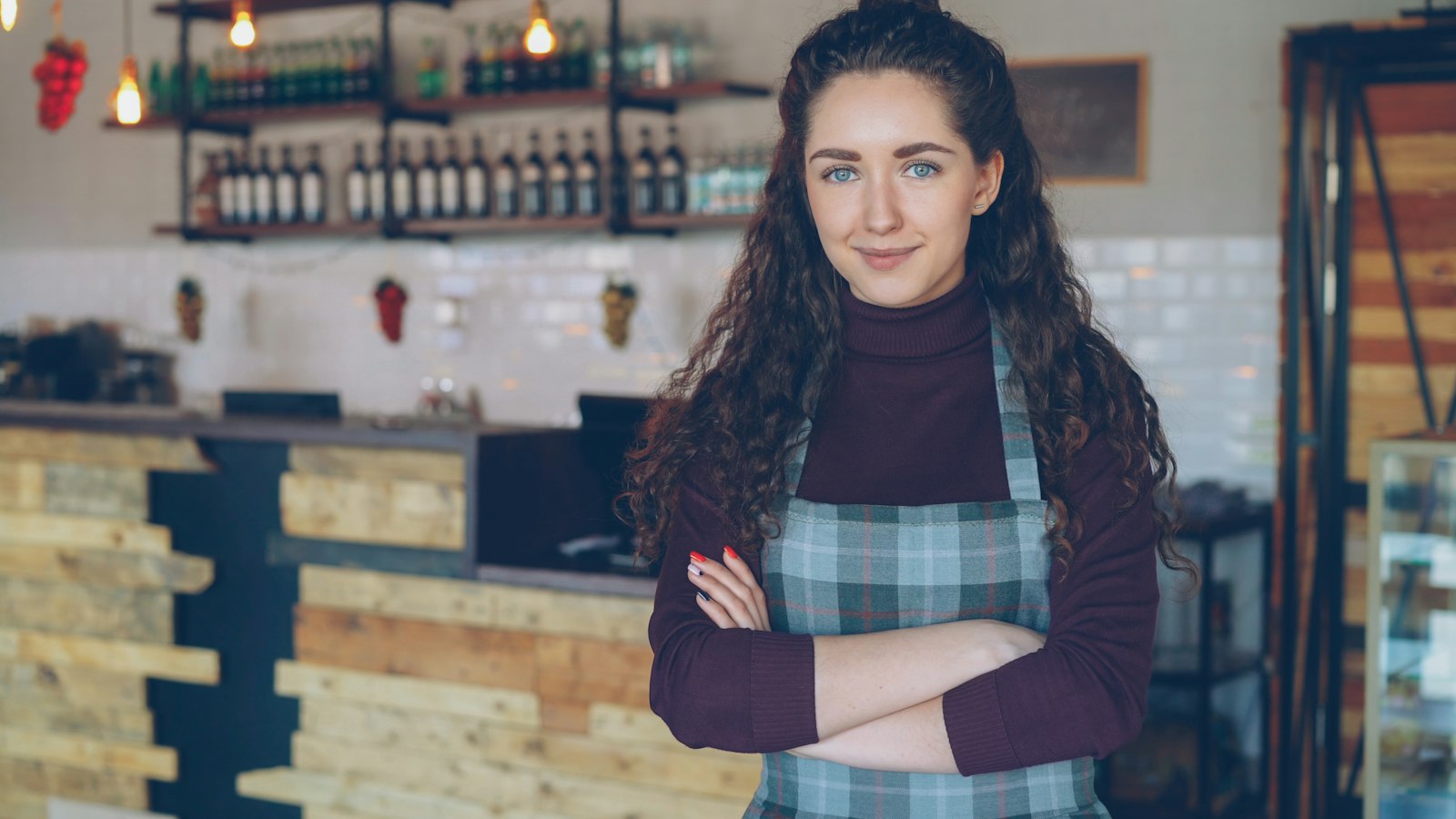 Owner of a small cafe in a plaid apron standing with arms folded in front of the counter and shelves of bottles